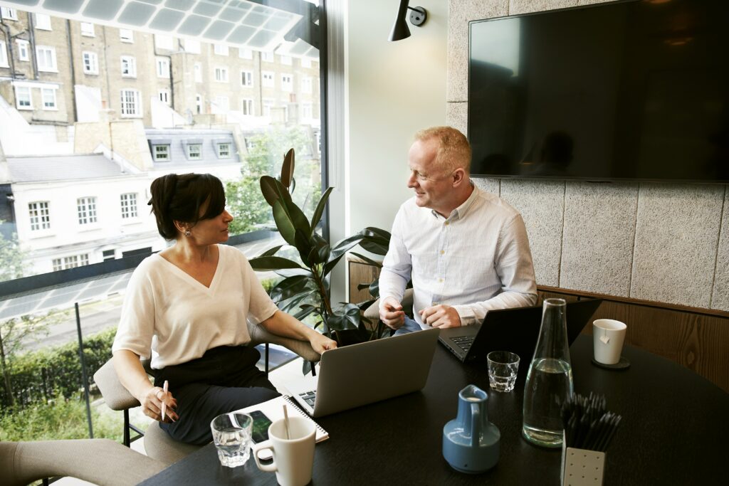 A man and a woman sat at a desk in an office, looking happy, calm with a laptop in front of them.